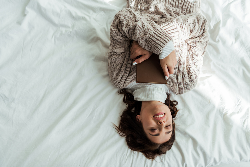 bovenaanzicht van lachende vrouw in trui liggend in bed in de ochtend  - Foto, afbeelding