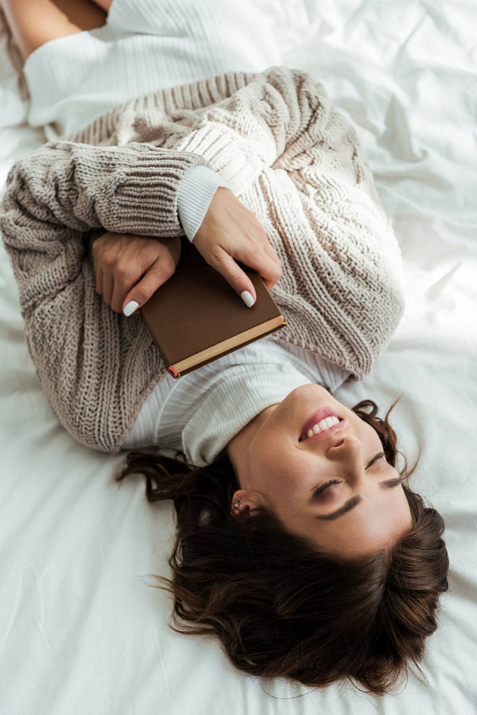 θέα υψηλής γωνίας χαμογελαστή γυναίκα σε πουλόβερ ξαπλωμένη στο κρεβάτι το πρωί  - Φωτογραφία, εικόνα