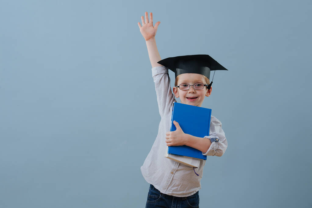 Zrzka první třídy chlapec v brýlích a studentský klobouk zvedá ruku nad modrou - Fotografie, Obrázek