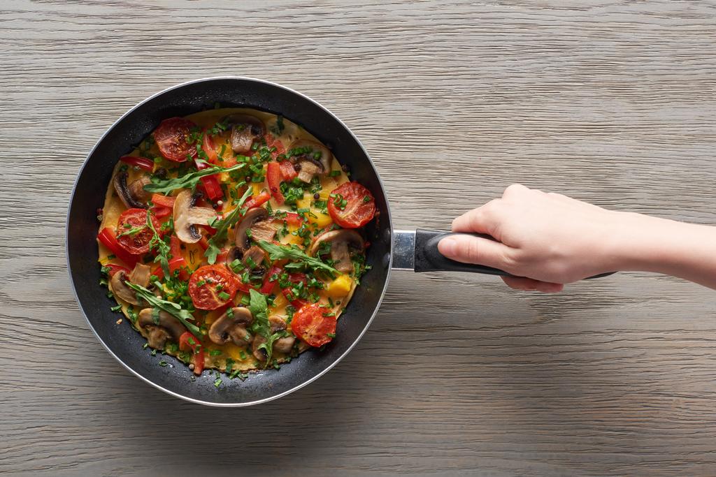 vista recortada de la mujer sosteniendo sartén con tortilla con setas, tomates y verduras
 - Foto, imagen