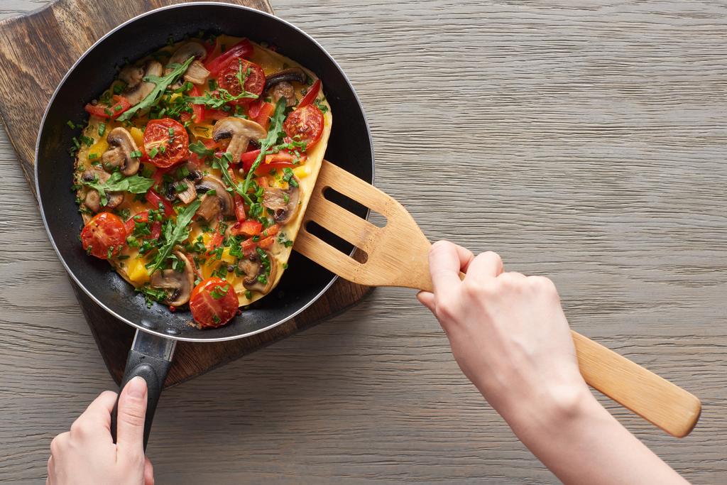 przycięty widok kobiety gotowanie omlet z grzybów, pomidorów i zieleni na patelni z drewnianą łopatą - Zdjęcie, obraz