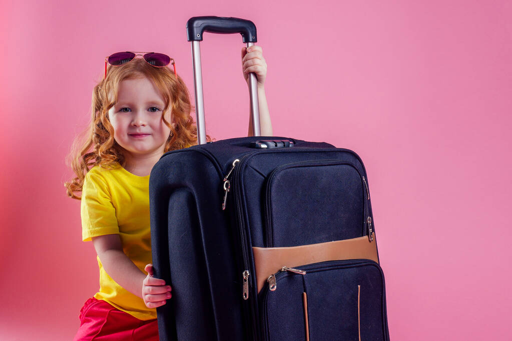 赤い髪の女性の子供ストライプ黄色のTシャツ大きな旅行スーツケースの上に座って夢は海の海岸ピンクの背景スタジオの残りの部分を飛ぶ - 写真・画像