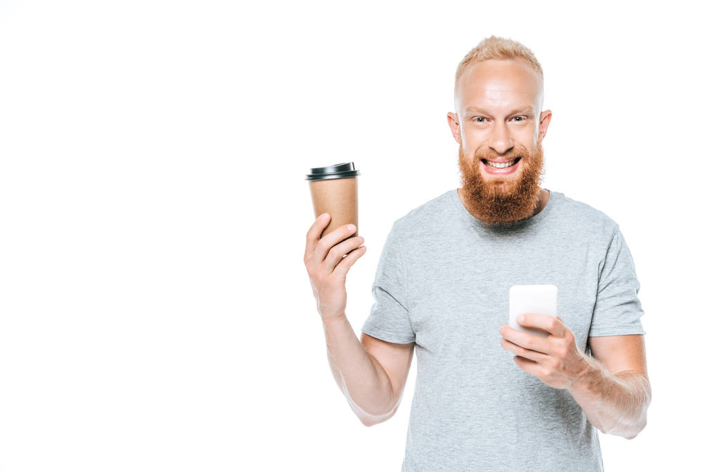 glimlachende man met baard en koffie te gaan met behulp van smartphone, geïsoleerd op wit - Foto, afbeelding