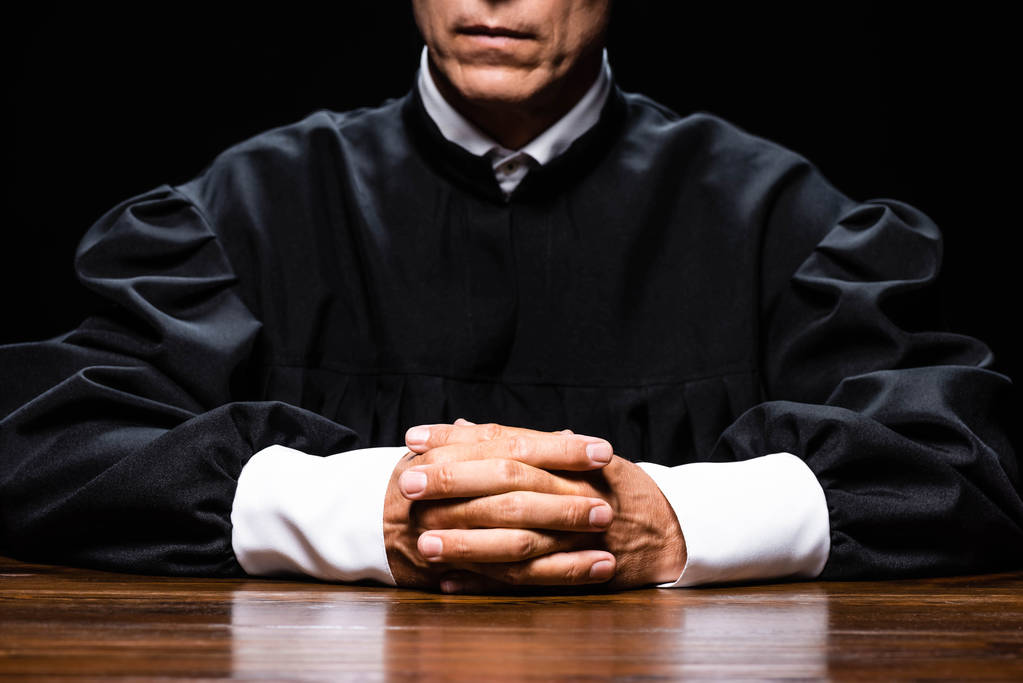 vista recortada del juez en bata judicial sentado a la mesa con las manos apretadas aisladas en negro
 - Foto, Imagen
