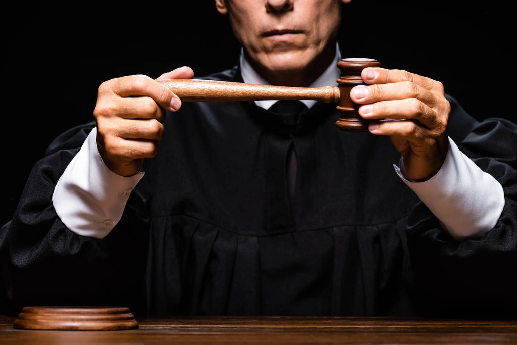 Ausgeschnittene Ansicht des Richters in Richterrobe, der am Tisch sitzt und den Hammer isoliert auf schwarz hält - Foto, Bild