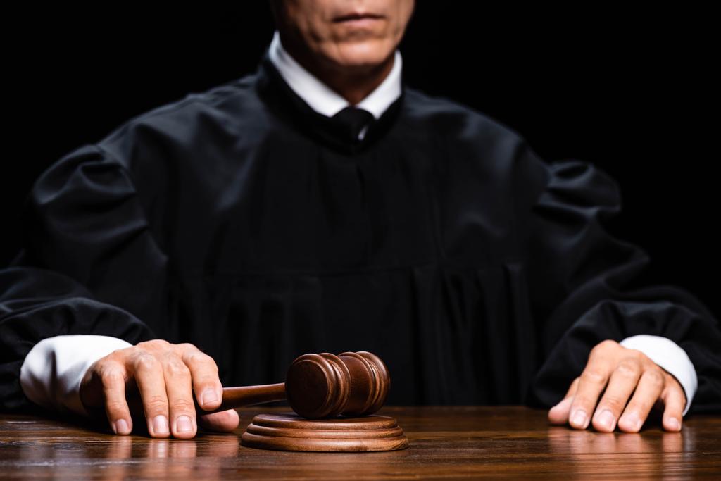 καλλιεργημένη άποψη του δικαστή με δικαστικό χιτώνα που κάθεται στο τραπέζι και κρατά το σφυρί απομονωμένο σε μαύρο - Φωτογραφία, εικόνα