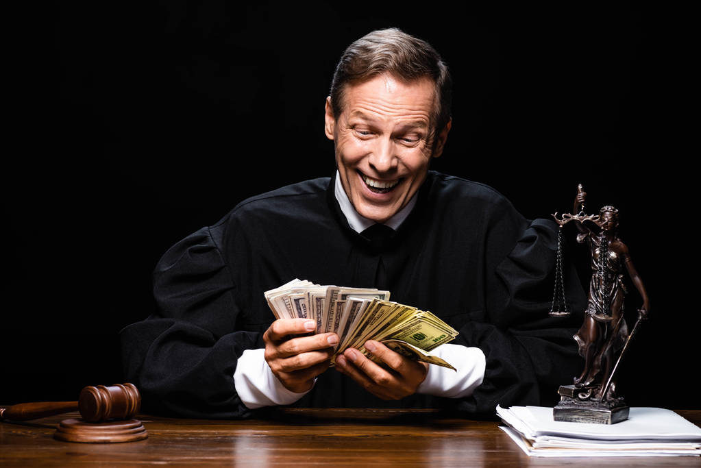 glimlachende rechter in gerechtelijk gewaad zit aan tafel en houdt dollarbankbiljetten geïsoleerd op zwart - Foto, afbeelding