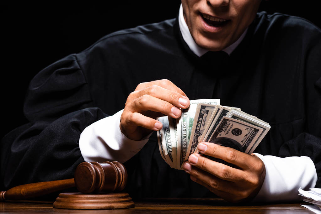 cropped view of shocked judge in judicial robe sitting at table and counting dollar banknotes isolated on black - Photo, Image