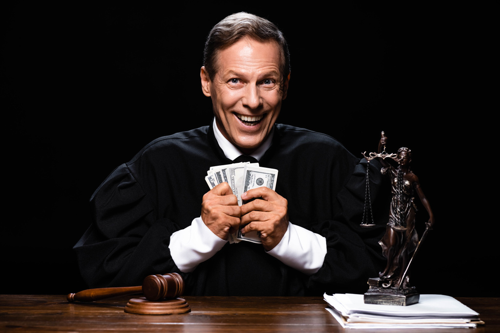 juge souriant en robe judiciaire assis à table et tenant des billets en dollars isolés sur noir
 - Photo, image