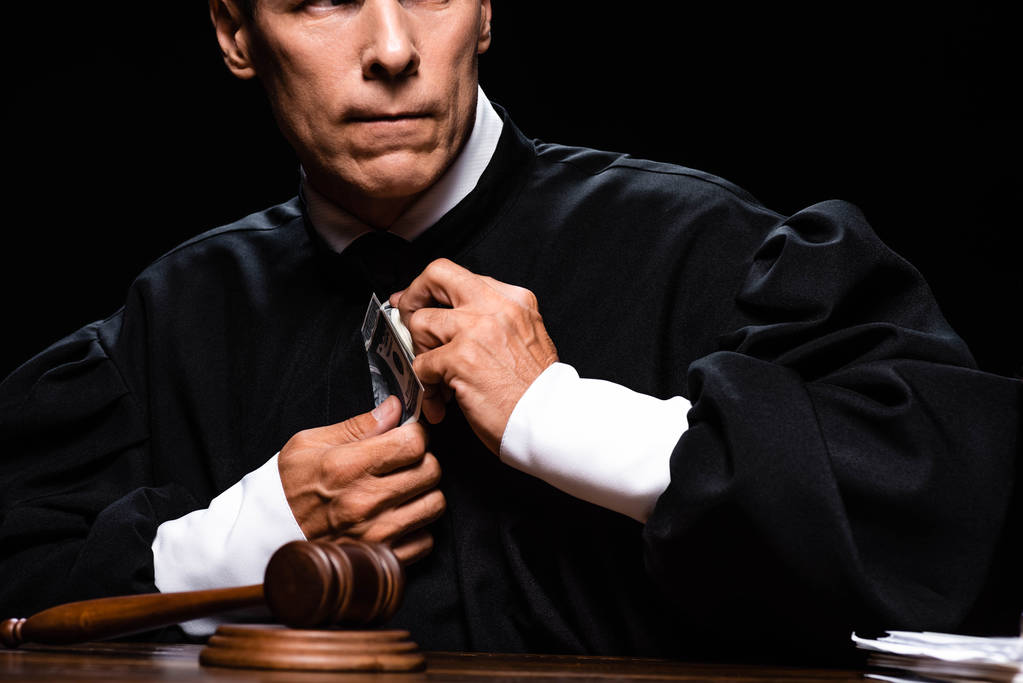 ostříhaný pohled vyděšeného soudce v soudní róbě sedí u stolu a drží dolarové bankovky izolované na černém - Fotografie, Obrázek