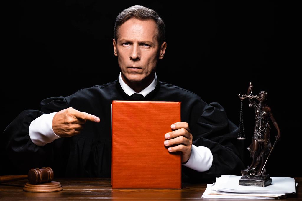δικαστής με δικαστική ρόμπα κάθεται στο τραπέζι και δείχνοντας με το δάχτυλο σε πορτοκαλί βιβλίο απομονώνονται σε μαύρο - Φωτογραφία, εικόνα