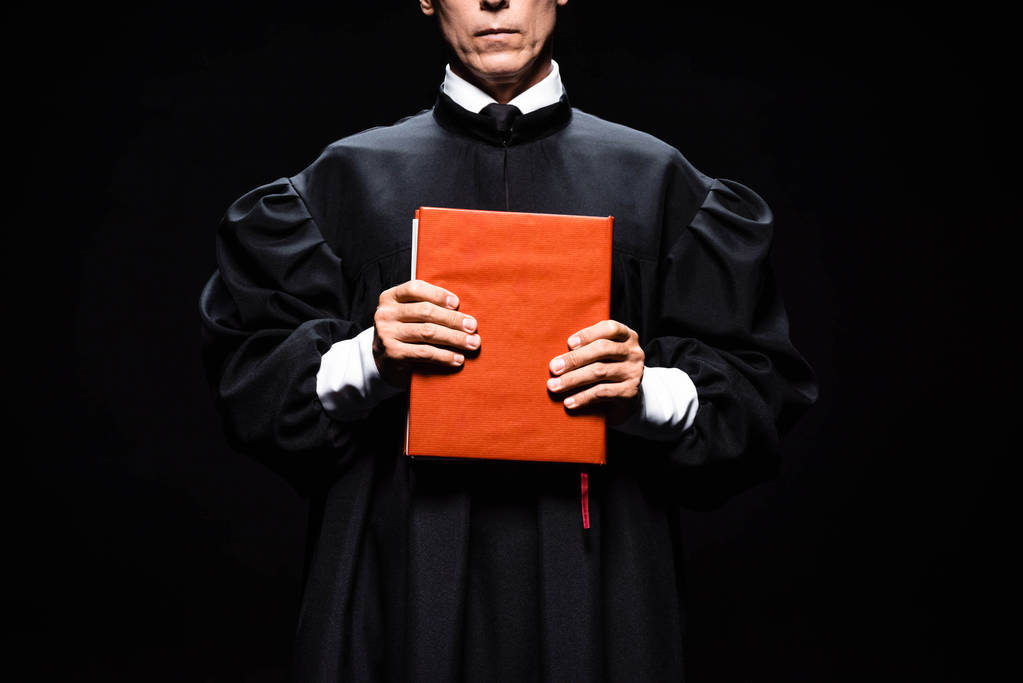 przycięty widok sędziego w szlafroku sądowym trzymającego pomarańczową książkę odizolowaną na czarno  - Zdjęcie, obraz