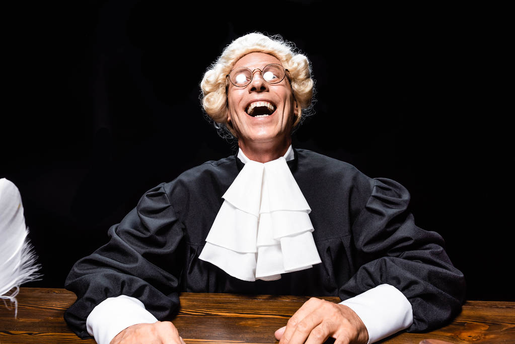 glimlachende rechter in gerechtelijk gewaad en pruik zitten aan tafel geïsoleerd op zwart - Foto, afbeelding