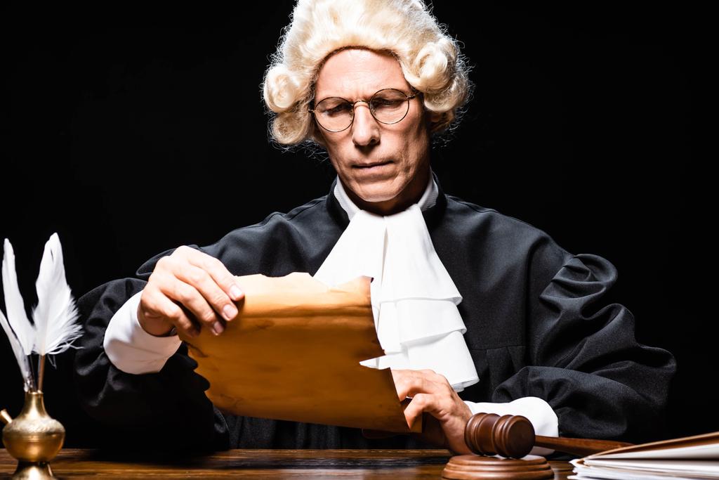 rechter in gerechtelijk gewaad en pruik zitten aan tafel en leesdocument geïsoleerd op zwart - Foto, afbeelding