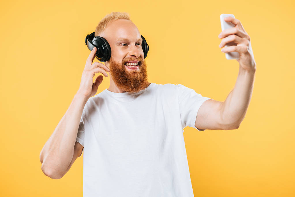 γενειάδα άνθρωπος ακούγοντας μουσική με ακουστικά και τη λήψη selfie στο smartphone, απομονώνονται σε κίτρινο - Φωτογραφία, εικόνα