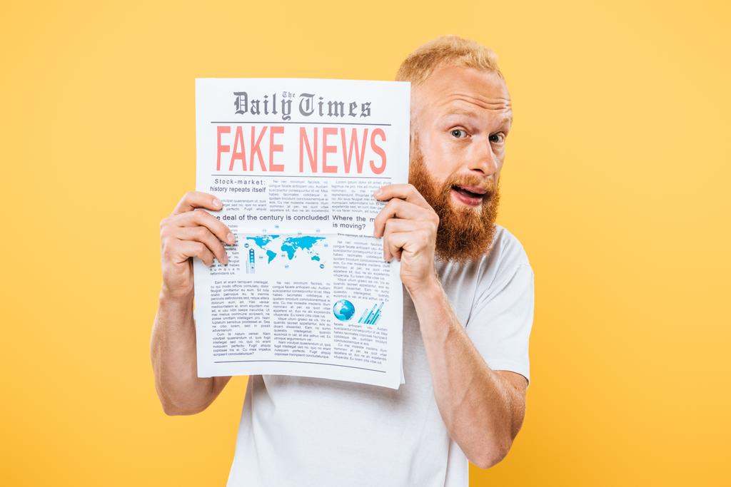 φοβισμένος γενειοφόρος άνδρας κρατώντας εφημερίδα με ψεύτικα νέα, απομονωμένος στο κίτρινο - Φωτογραφία, εικόνα