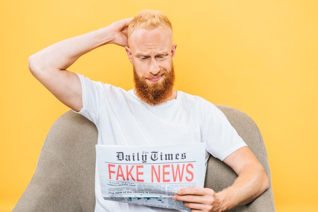 вдумчивый бородатый мужчина читает газету с фальшивыми новостями, сидя на кресле, изолированном на желтом
 - Фото, изображение