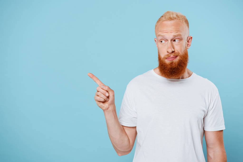 portret van sarcastische man met baard in wit t-shirt wijzend naar iets, geïsoleerd op blauw  - Foto, afbeelding