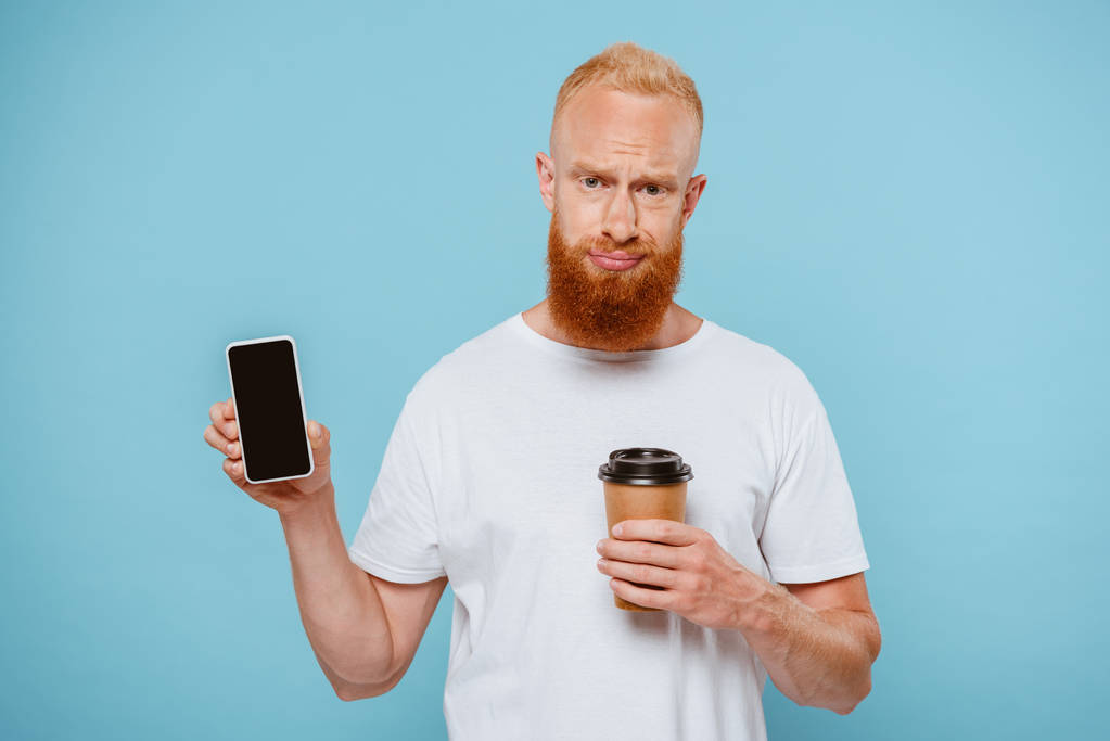 szkeptikus szakállas férfi kávéval menni bemutató okostelefon üres képernyőn, elszigetelt kék - Fotó, kép