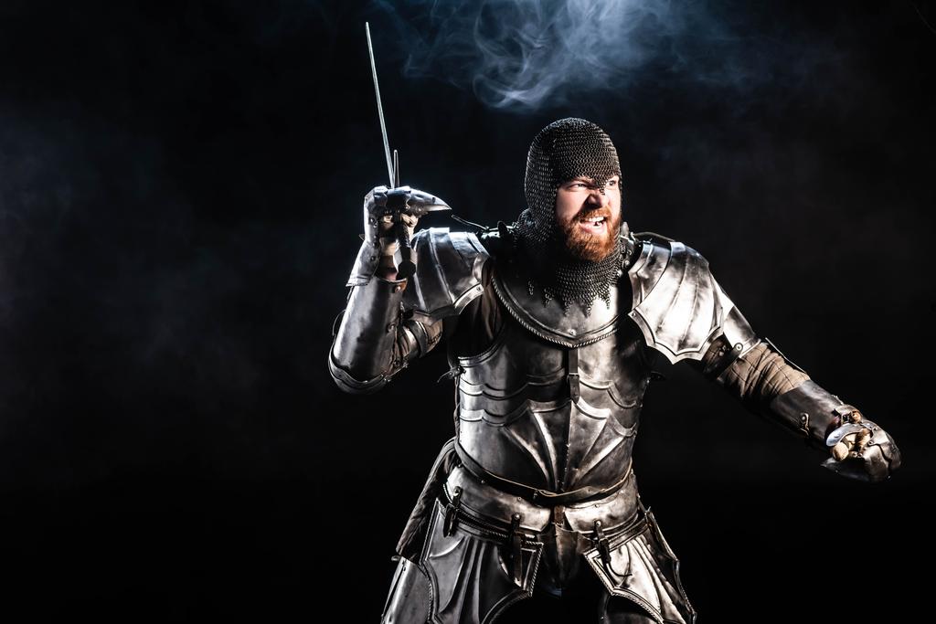 όμορφος και θυμωμένος ιππότης με πανοπλία πολεμώντας με σπαθί σε μαύρο φόντο  - Φωτογραφία, εικόνα