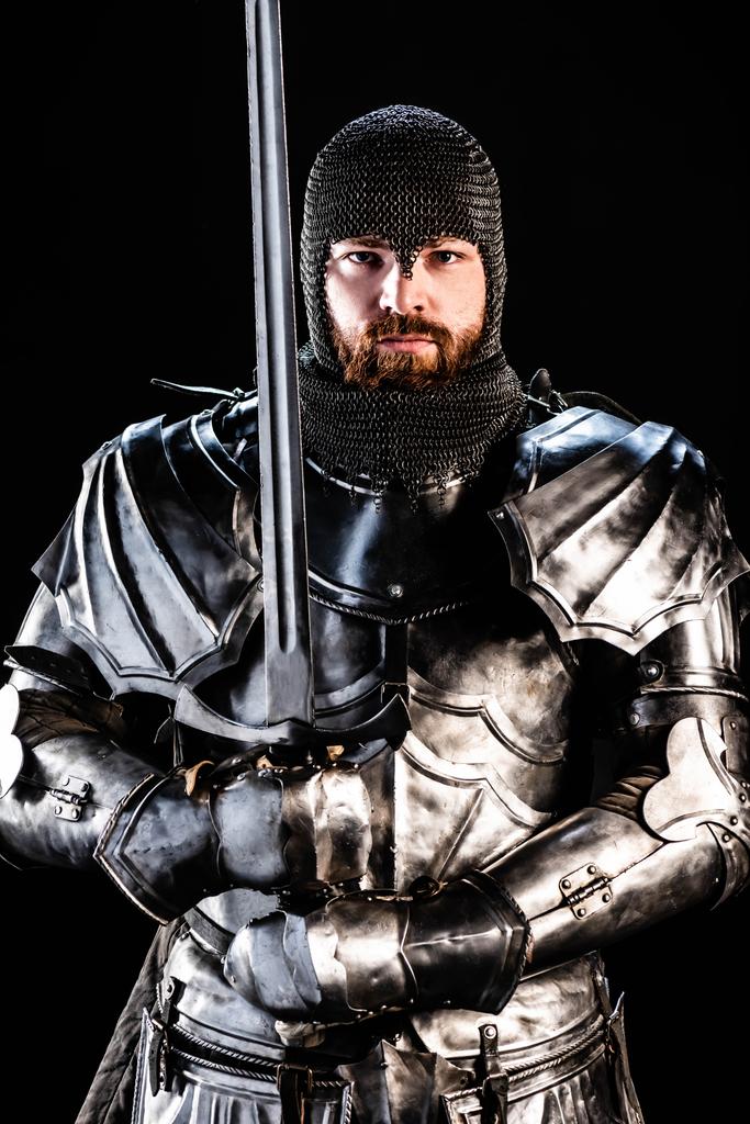 handsome knight in armor looking at camera and holding sword isolated on black - Photo, Image