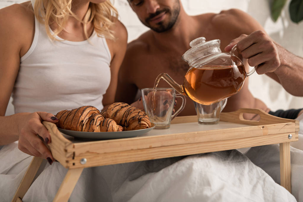 kivágott kilátás fiatal pár tea és croissant tálcán az ágyban reggel   - Fotó, kép