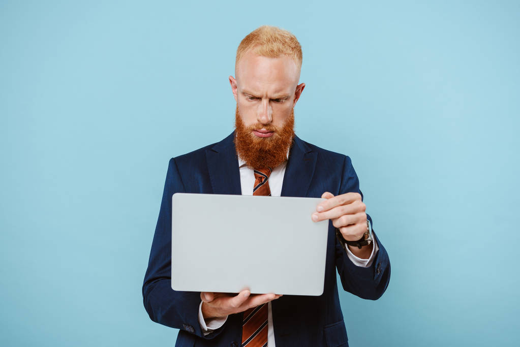 серьезный бородатый бизнесмен в костюме с ноутбуком, изолированный на синий
 - Фото, изображение