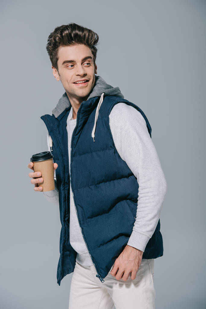 styling χαμογελαστός άνθρωπος σε ζεστό γιλέκο κρατώντας καφέ για να πάει, απομονώνονται σε γκρι - Φωτογραφία, εικόνα
