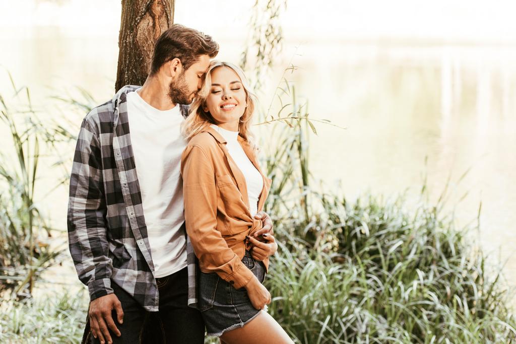 young man embracing cheerful girlfriend while standing near lake in park - Photo, Image