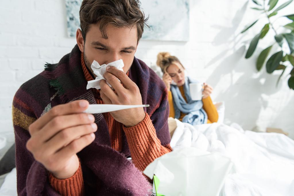 άρρωστος άνθρωπος με πυρετό κρατώντας θερμόμετρο και χαρτοπετσέτα στο υπνοδωμάτιο με γυναίκα πίσω - Φωτογραφία, εικόνα