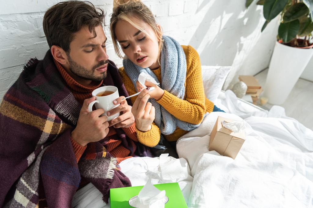 casal doente com febre segurando termômetro, bebida quente e guardanapos na cama
 - Foto, Imagem
