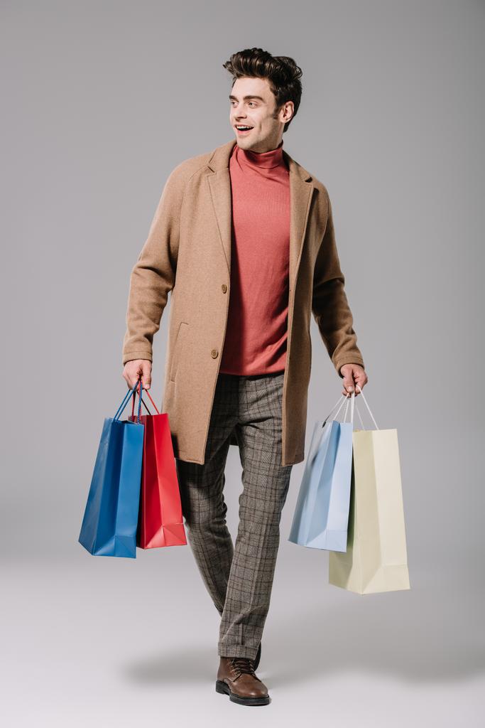 happy fashionable man in beige coat walking with shopping bags on grey - Photo, Image