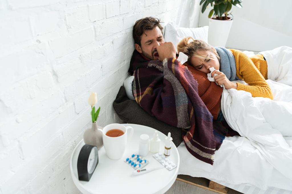 ill couple holding napkins in bedroom with thermometer, pills, hot drink and nasal spray - Zdjęcie, obraz