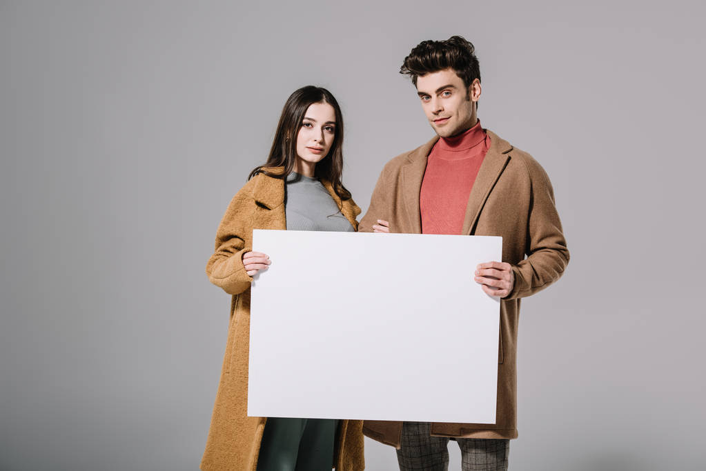 fashionable couple in beige coats posing with empty board, isolated on grey - Photo, Image