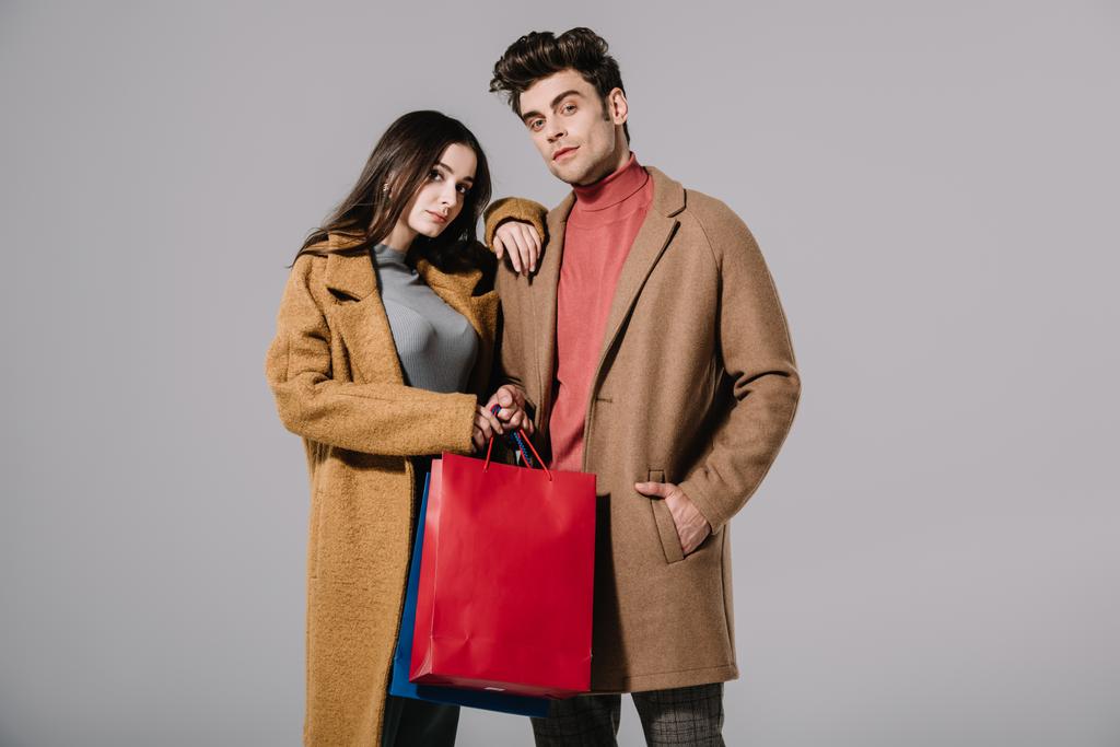 Gri renkte izole edilmiş alışveriş torbalarıyla poz veren bej ceketli şık bir çift. - Fotoğraf, Görsel