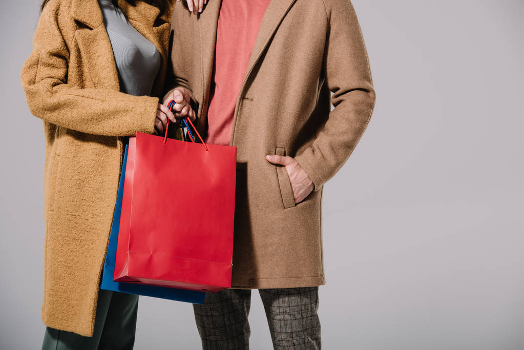 bijgesneden beeld van paar in beige jassen houden boodschappentassen geïsoleerd op grijs - Foto, afbeelding