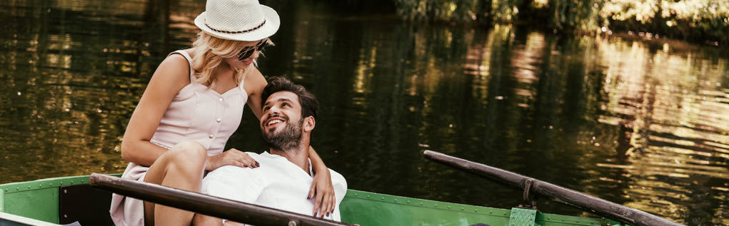 panoramische Aufnahme einer jungen Frau in Anzug und Hut, die ihren glücklichen Freund umarmt, während sie im Boot sitzt - Foto, Bild