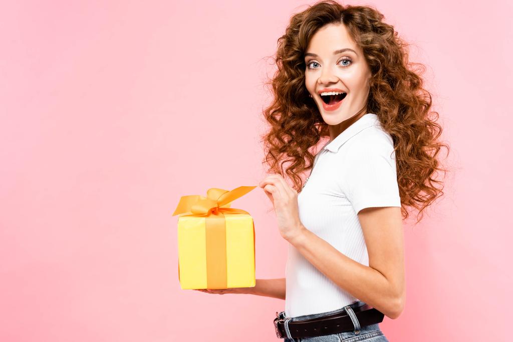excited attractive curly girl holding gift box, isolated on pink - 写真・画像