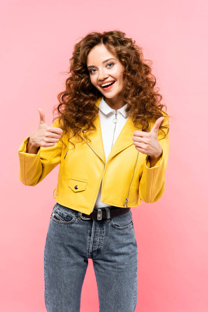 attractive curly girl in yellow leather jacket showing thumbs up, isolated on pink - 写真・画像