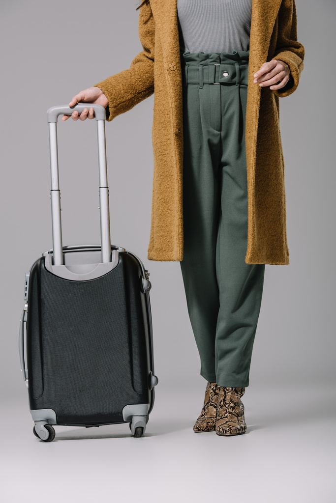 обрізаний вид стильної жінки в бежевому пальто позує з дорожньою сумкою на сірому
 - Фото, зображення