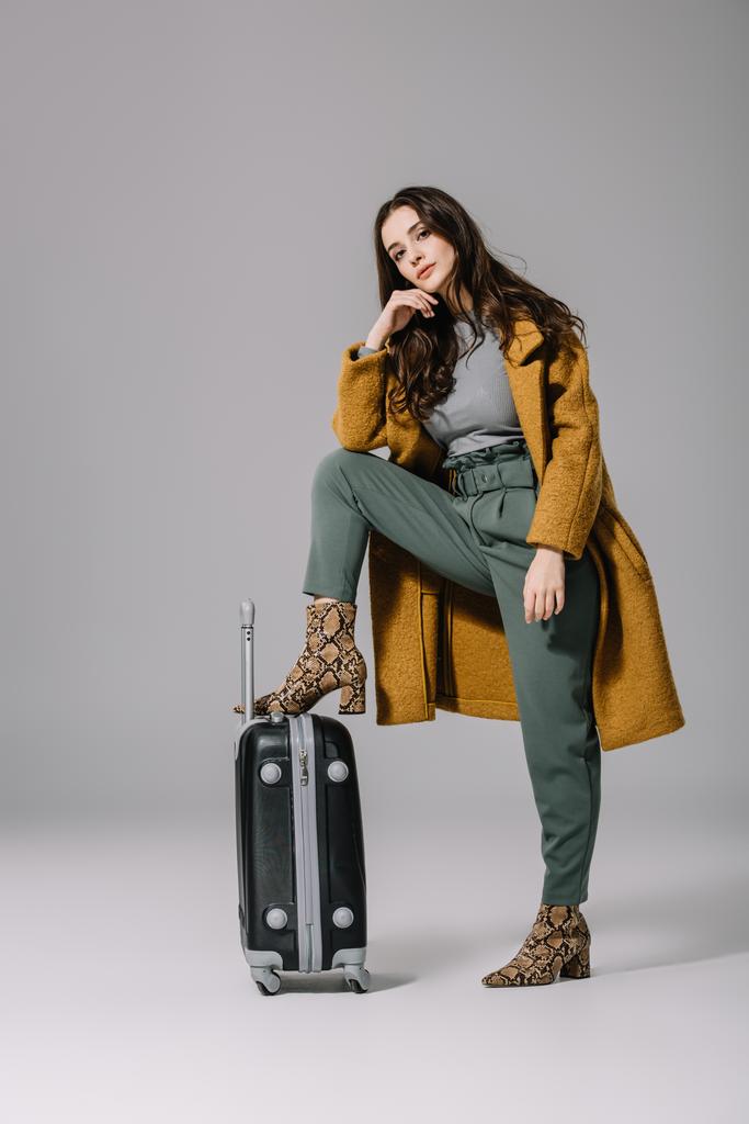 fashionable woman in beige coat posing with travel bag on grey - Photo, Image