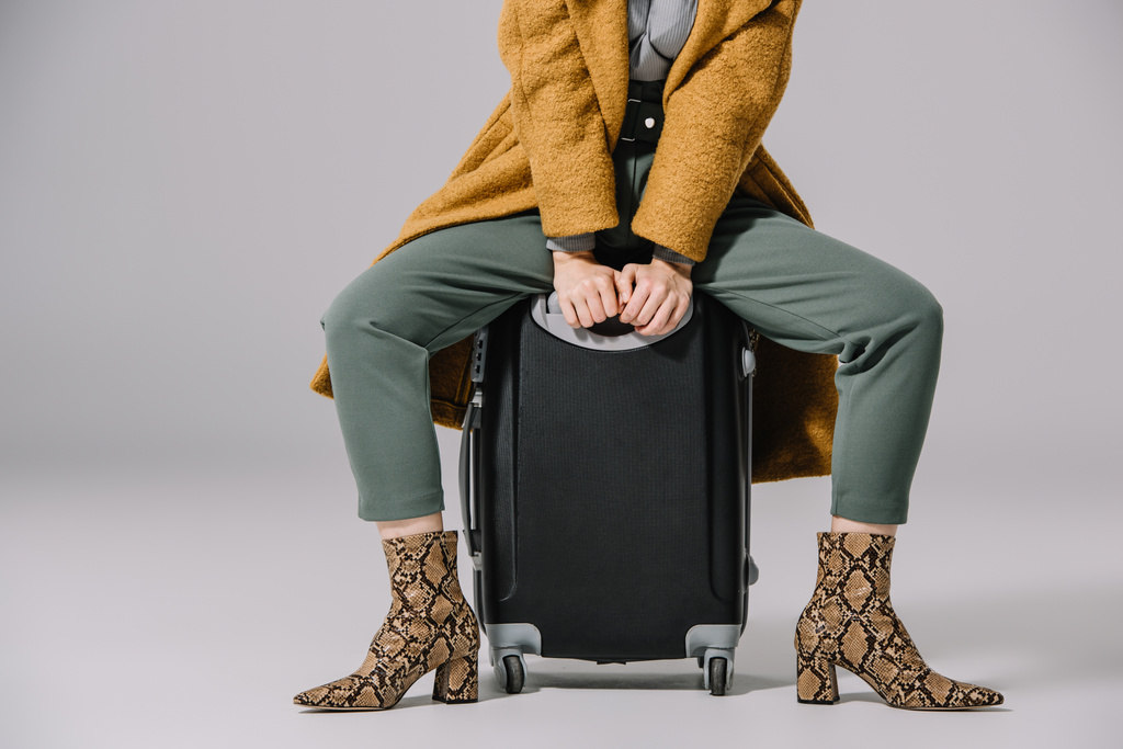 przycięty widok stylowej kobiety w beżowym płaszczu siedzi na torbie podróżnej na szarym - Zdjęcie, obraz