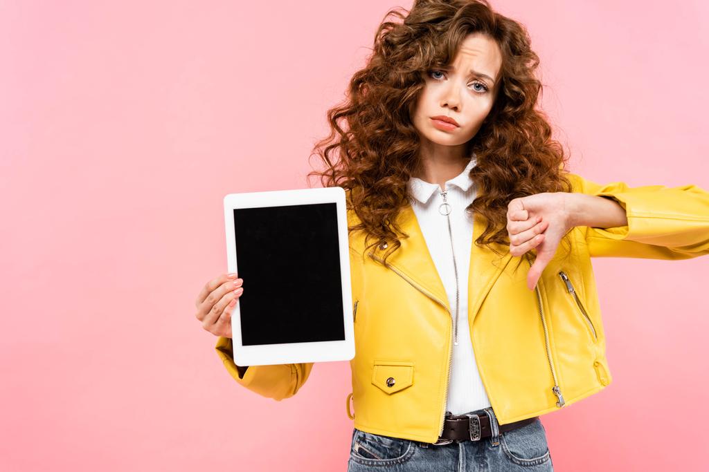 smutná kudrnatá dívka ukazující palec dolů a digitální tablet s prázdnou obrazovkou, izolované na růžové - Fotografie, Obrázek