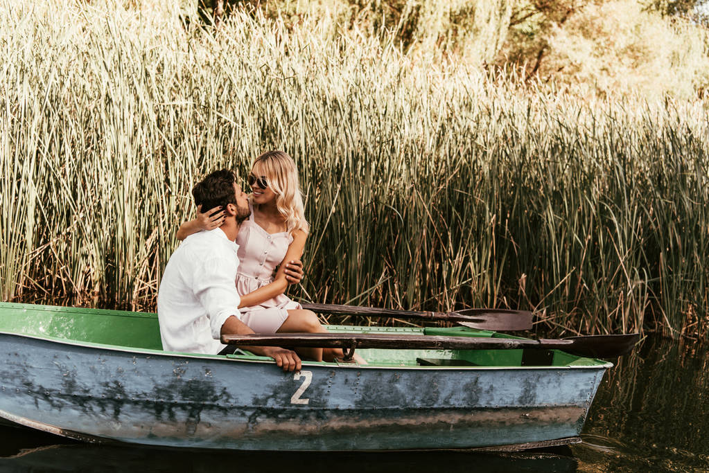 νεαρό ζευγάρι που αγκαλιάζεται και φιλιέται σε βάρκα στο ποτάμι κοντά σε θάμνους - Φωτογραφία, εικόνα