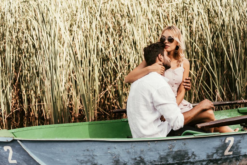 aantrekkelijke jonge vrouw omarmen vriendje in boot op meer in de buurt struikgewas van sedge - Foto, afbeelding