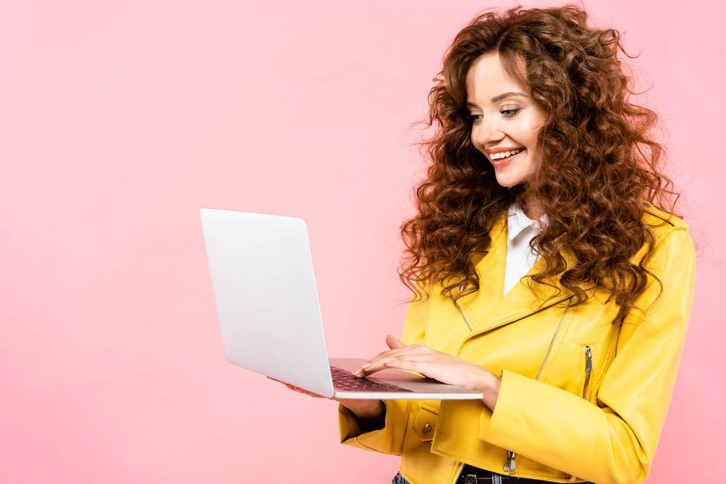 gelukkig krullend vrouw met behulp van laptop, geïsoleerd op roze - Foto, afbeelding