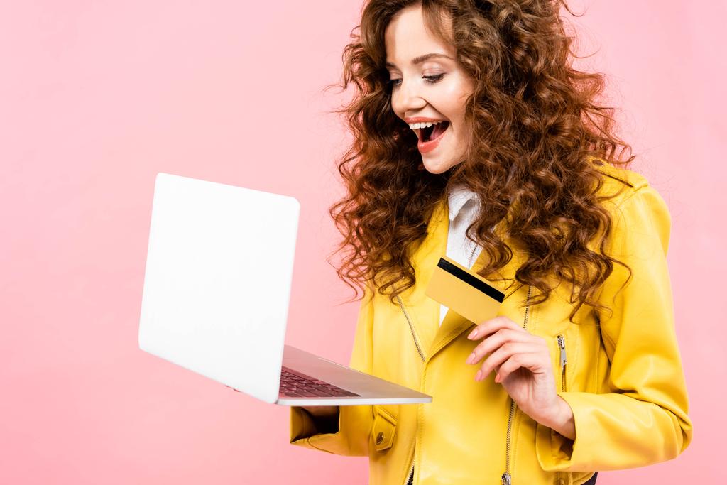 vzrušený kudrnatý žena nakupování on-line s kreditní kartou a notebookem, izolované na růžové - Fotografie, Obrázek