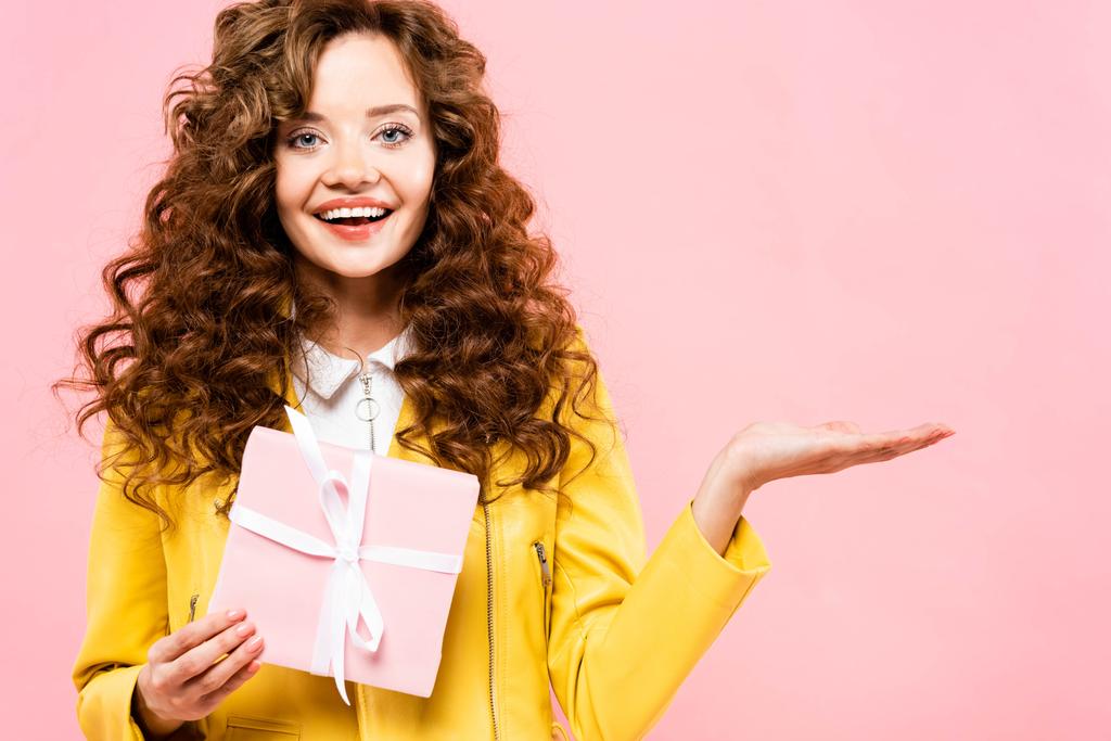 όμορφο σγουρό κορίτσι κρατώντας δώρο και παρουσιάζοντας κάτι, απομονωμένο σε ροζ - Φωτογραφία, εικόνα