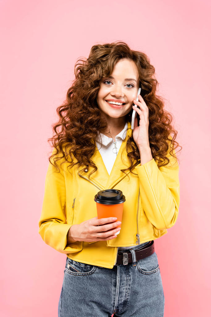 cheerful girl with disposable cup of coffee talking on smartphone, isolated on pink - Photo, Image