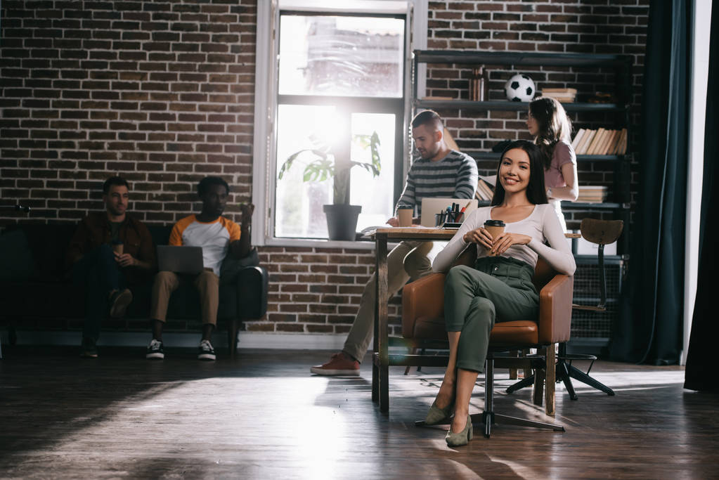привлекательная деловая женщина, сидящая в кресле с кофе, чтобы пойти и улыбаясь в камеру возле мультикультурных коллег
 - Фото, изображение